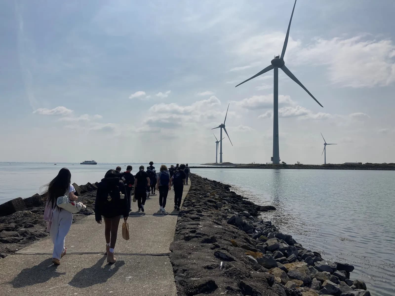 A group of people walk along a paved path next to a body of water, with large wind turbines visible in the distance under a partly cloudy sky.